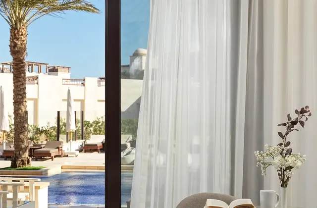 Bedroom nook with sheer curtains, an open book and flowers on a table beside a pool view window