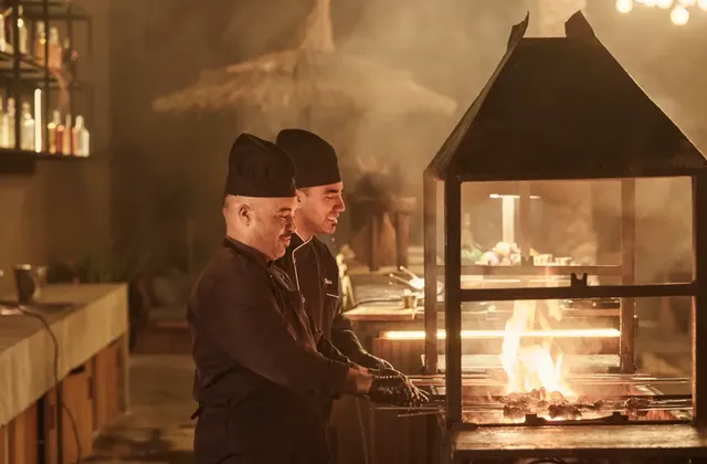 Two chefs cooking meat over an open flame grill in a dimly lit restaurant kitchen