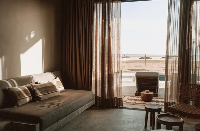 Living room with a sofa and sheer curtains opening onto a beach view with loungers