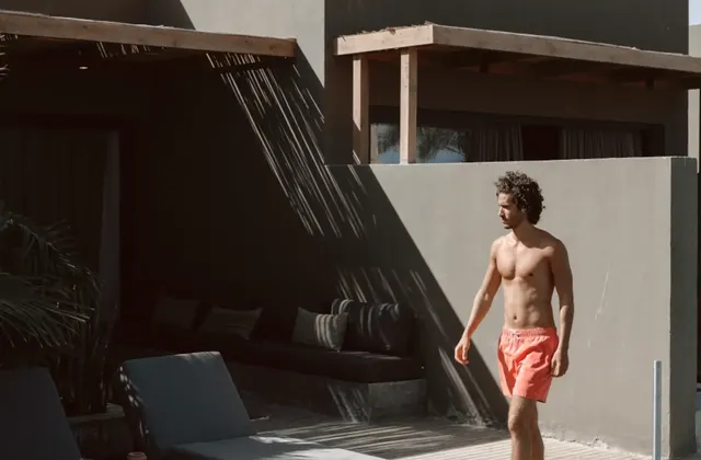 Man in red swim shorts walking along a poolside deck past dark loungers and a shaded lounge area