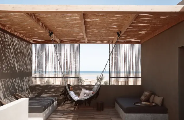 Covered terrace with a hanging hammock chair and lounge seating, framing a view of the beach and sea
