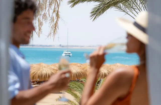 Couple toasting with champagne glasses on a beach terrace with the sea and thatched umbrellas in view