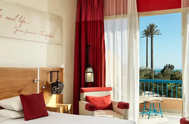 Hotel room with a red-and-white bed, red curtains and a balcony overlooking palm trees and the sea