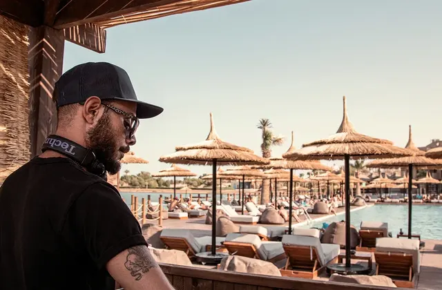 DJ performing by the pool at Cooks Club El Gouna, setting a lively and energetic atmosphere. The DJ booth is positioned near the poolside, with speakers and equipment arranged for optimal sound. Guests can be seen relaxing on lounge chairs or swimming, enjoying the vibrant music. The scene is enhanced by sunny weather and clear blue skies, creating a perfect backdrop for a fun and social poolside experience.