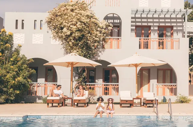 Guests relaxing on loungers under umbrellas beside a pool in front of a white arched hotel building