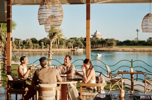 Group of friends dining at a waterside table under woven pendant lamps, a lagoon and palm trees behind them