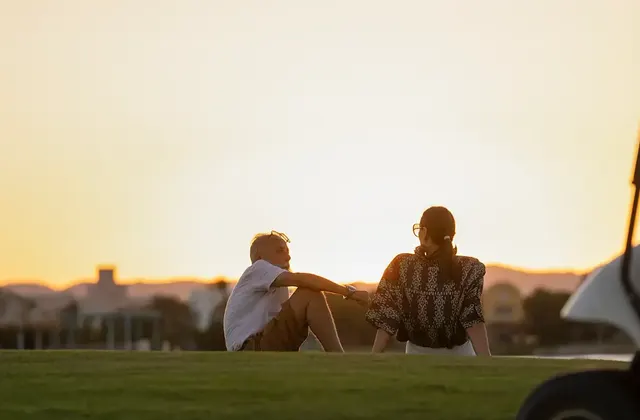Two people sitting on grass watching the sunset, a golf cart parked beside them