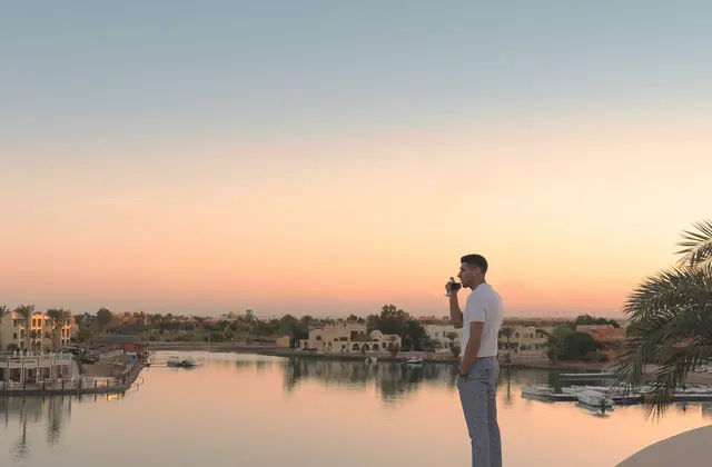 Man standing on a waterfront ledge drinking coffee at sunset with a lagoon and buildings behind him
