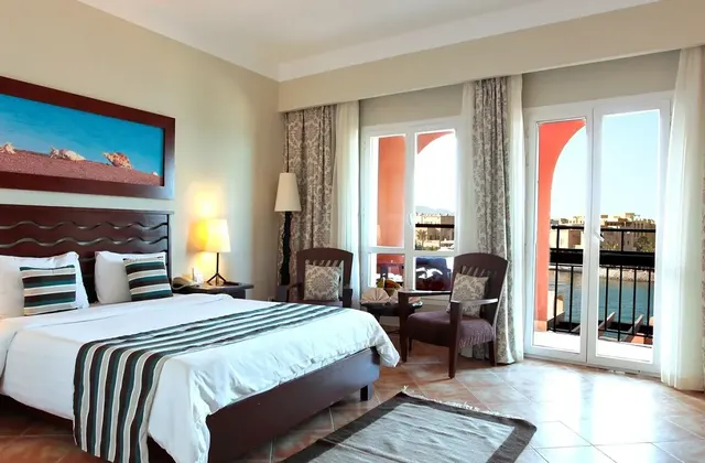 Ocean view standard room at Three Corners - Ocean View, featuring an open curtain balcony with a breathtaking view of the sea. The room includes a large bed, two chairs, and a painting on the wall, creating a cozy and artistic atmosphere.